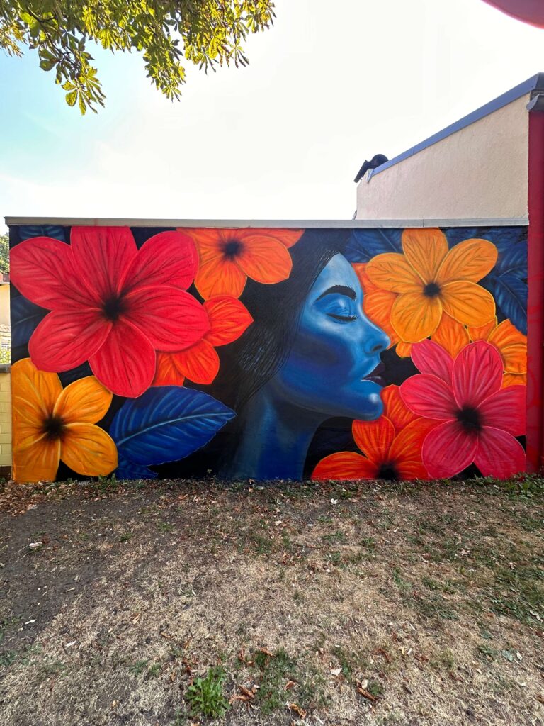 Graffiti von Graugegner: blaues Frauenprofil zwischen großen roten und orangefarbenen Blüten auf einer Außenwand.