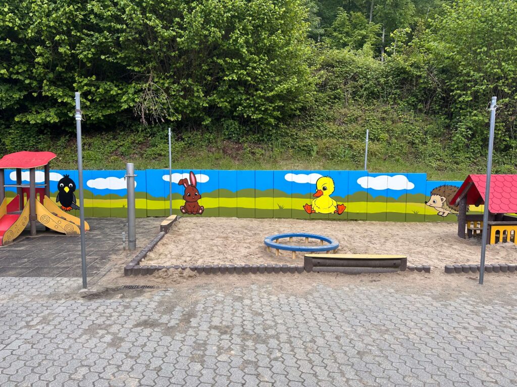 Graffiti von Graugegner an einer Spielplatzwand mit Vogel, Hase, Ente und Igel vor einer grünen Landschaft und blauem Himmel.